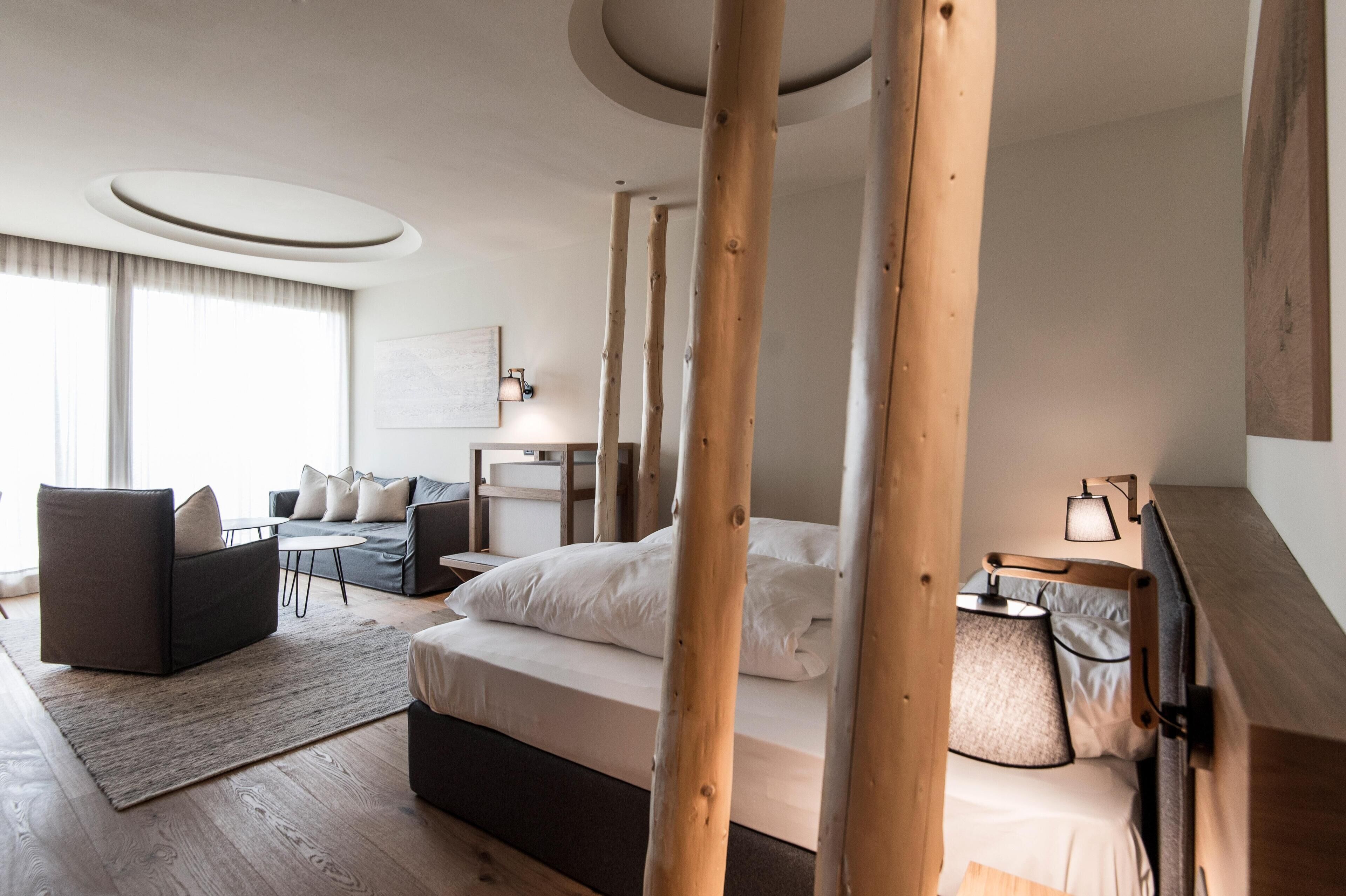 A modern hotel room with a large bed, a sofa, a small table, and a desk. There are wooden beams in the ceiling and a large window with curtains.