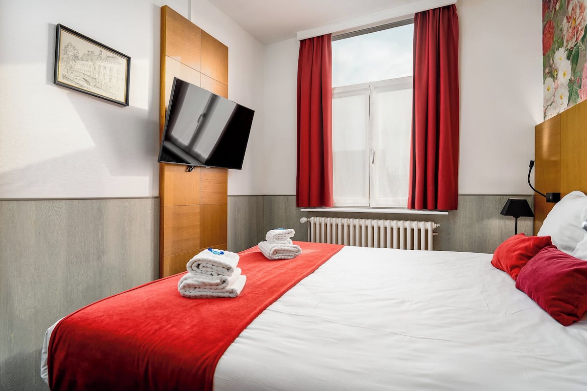 A hotel room with a bed, red curtains, a mounted TV, and a framed picture on the wall.