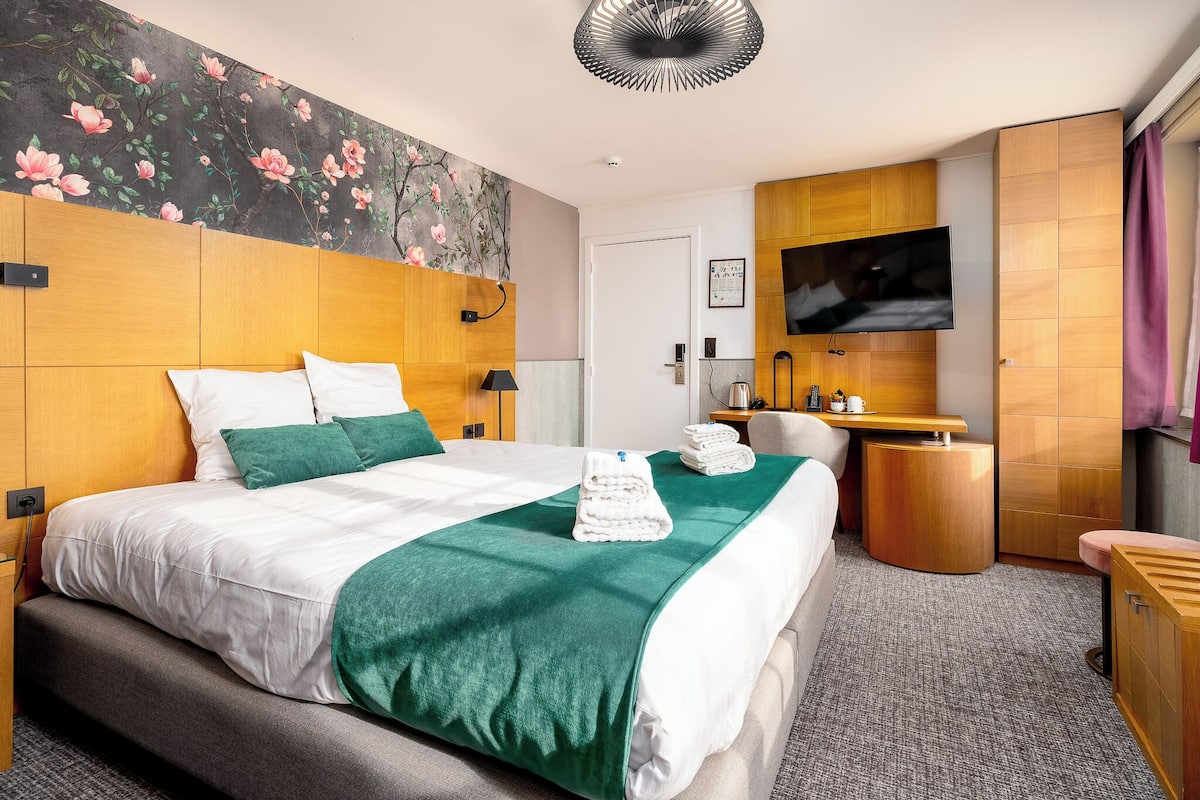 A hotel room with a large bed, a TV mounted on the wall, a wooden headboard, and a floral mural on the wall.
