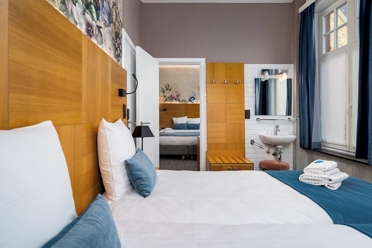 A hotel room with a wooden headboard, a bed with white and blue bedding, a bedside table with a lamp, a small wooden stool, a bathroom with a sink and mirror, and a window with curtains.