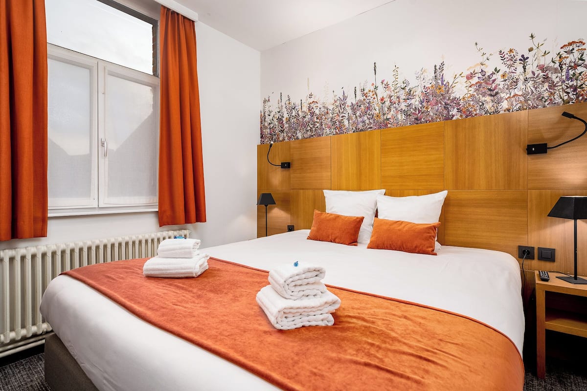 A hotel room with a large bed, orange curtains, a floral wall mural, and a wooden headboard.