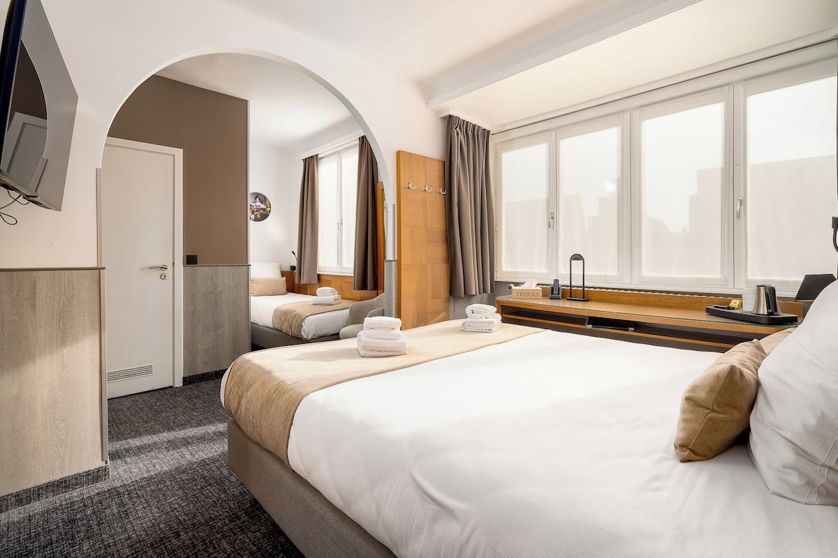 A hotel room with a large bed, a desk with a lamp, a TV mounted on the wall, and a window with curtains.