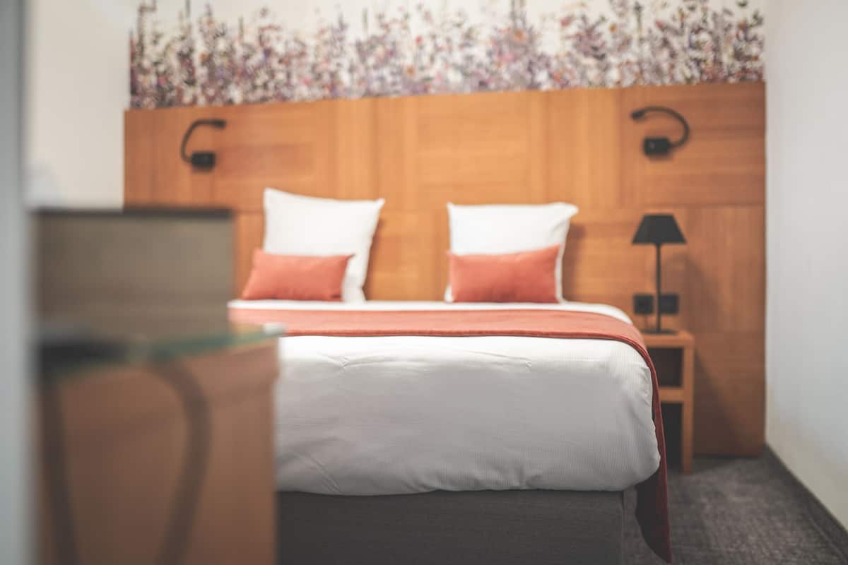 A bedroom with a wooden headboard, two pillows, a bedside table with a lamp, and a floral wall decoration.