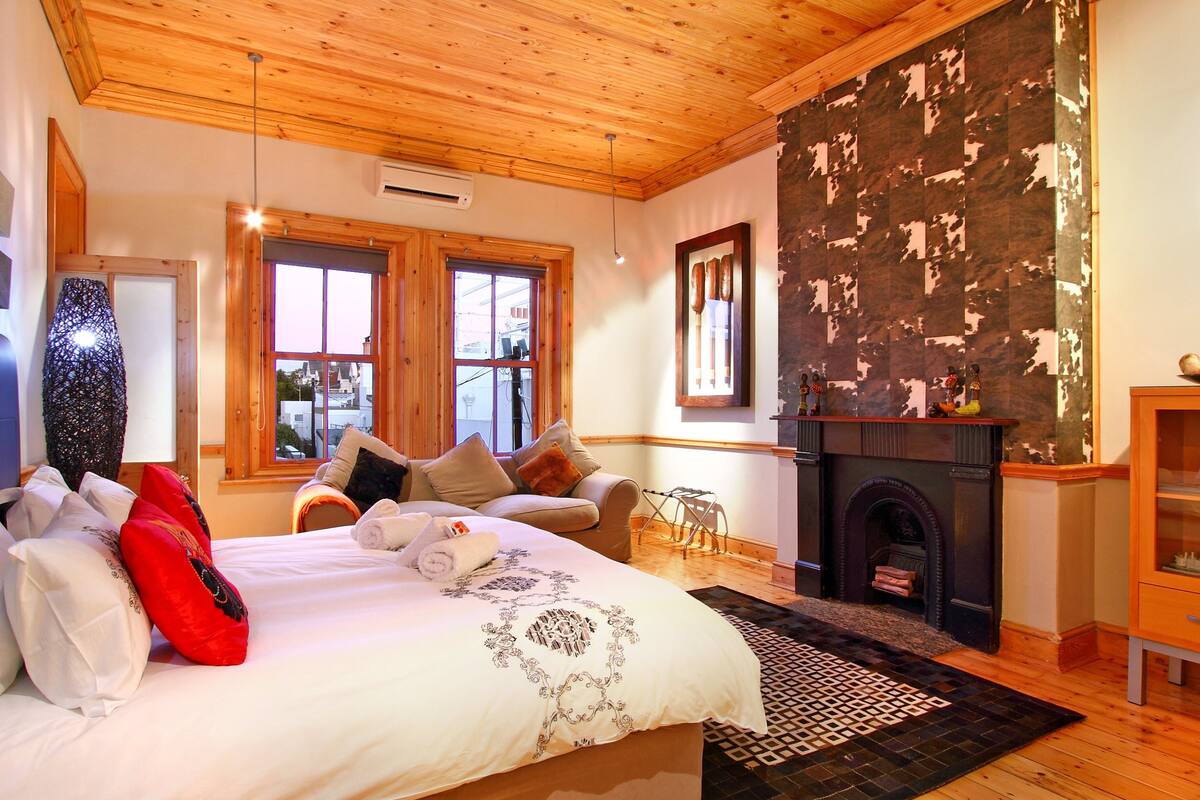 A bedroom with a wooden ceiling, a fireplace, a bed with pillows, and a window.