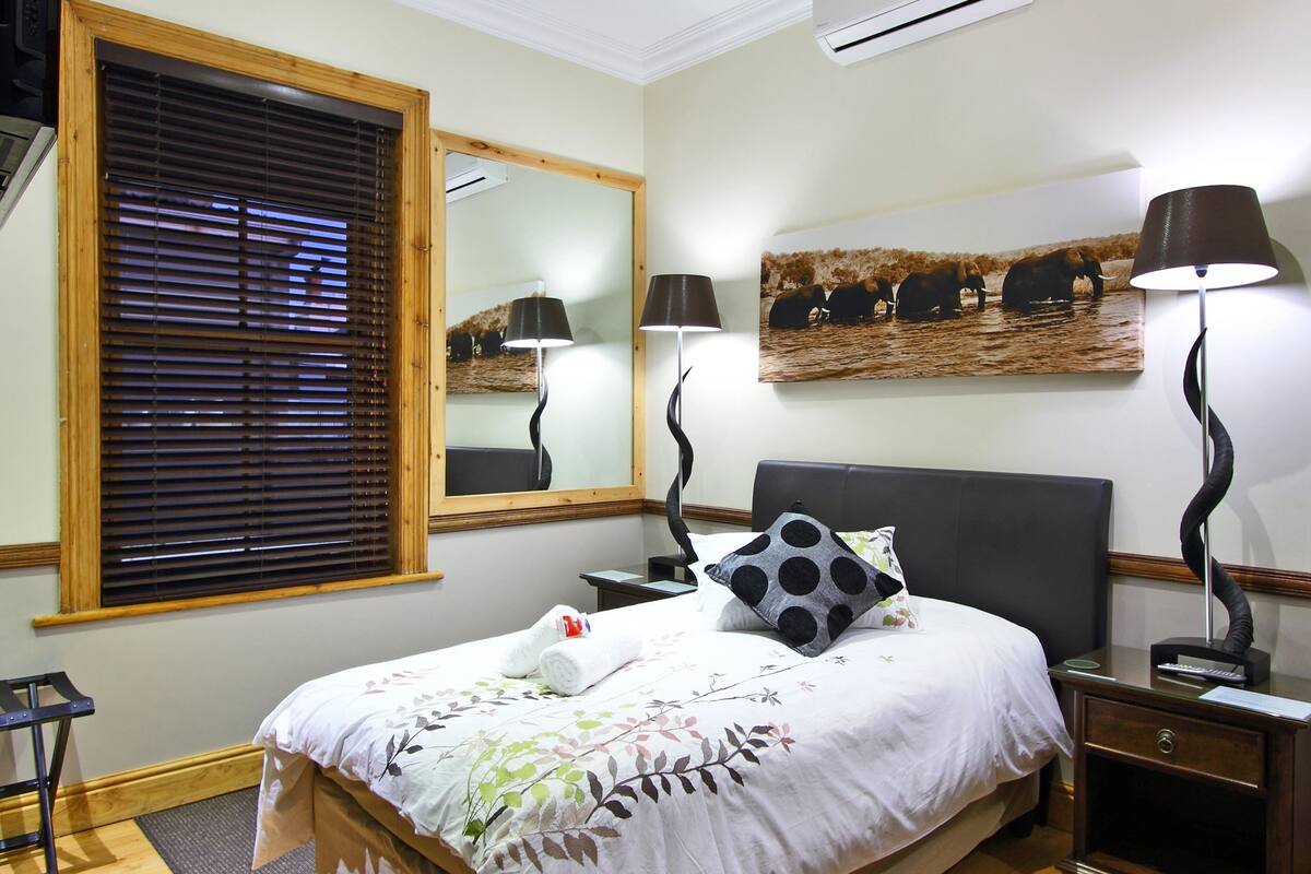 A neatly arranged hotel room with a bed, a nightstand, a lamp, a mirror, and a painting of elephants.