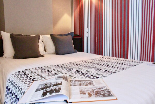 Una cama bien tendida con una colcha de estampado geométrico, un libro abierto sobre la cama y una cortina a rayas al fondo.