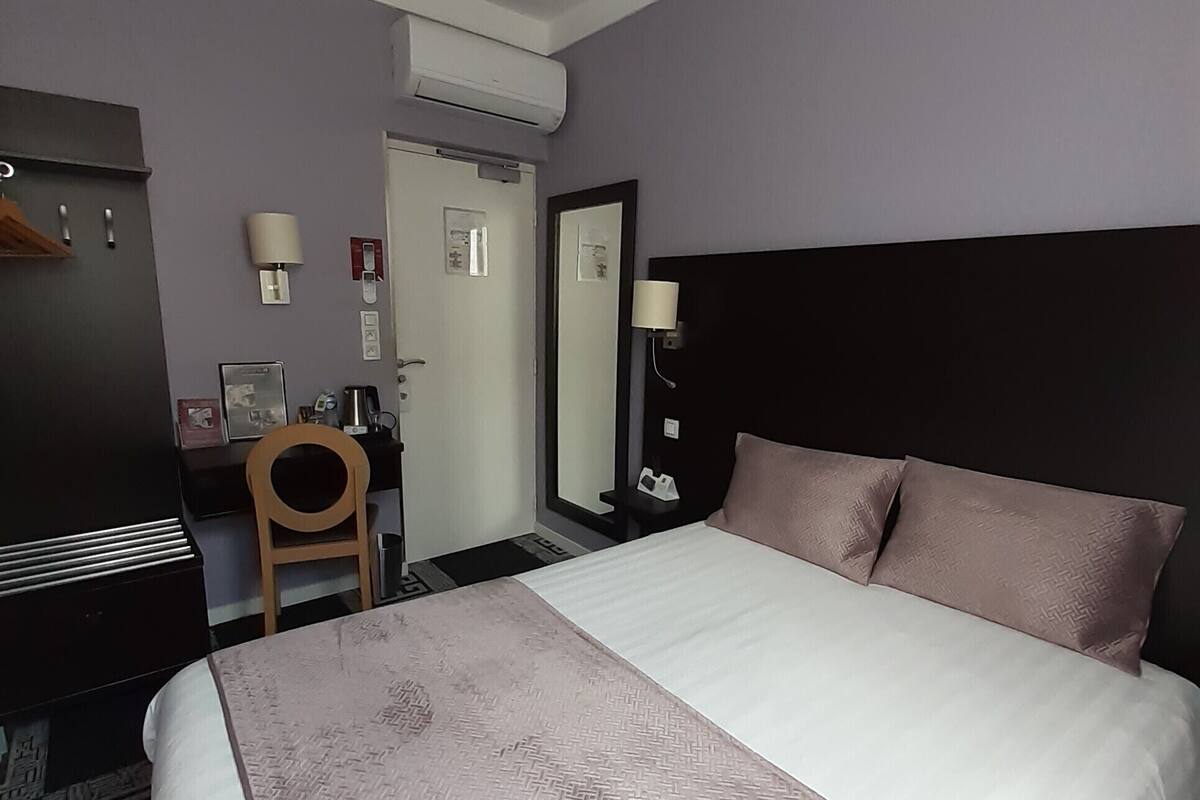 A hotel room with a large bed, a desk, a chair, a wardrobe, and a mirror.