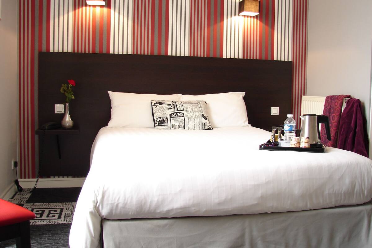 A hotel room with a large bed, a nightstand, a vase with flowers, a tray with a kettle, and a red chair.