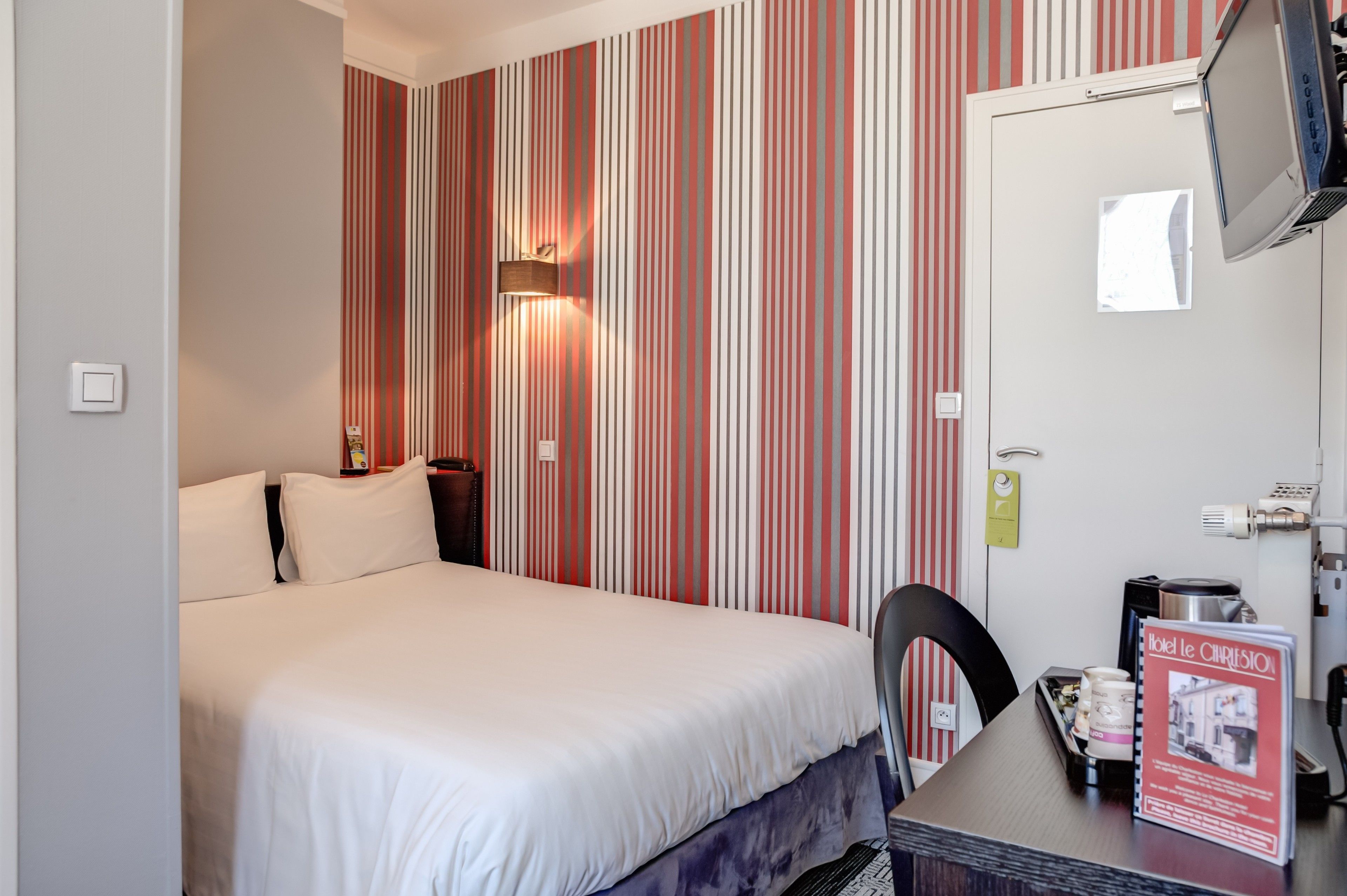 Quarto de hotel com cama, escrivaninha com cadeira, televisão na parede e papel de parede listrado.