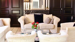 Ein Wohnzimmer mit einem beigen Sofa, zwei Sesseln, einem Couchtisch mit einer Vase voller roter Blumen, einem Kamin und dunklen Holzvertäfelungen.