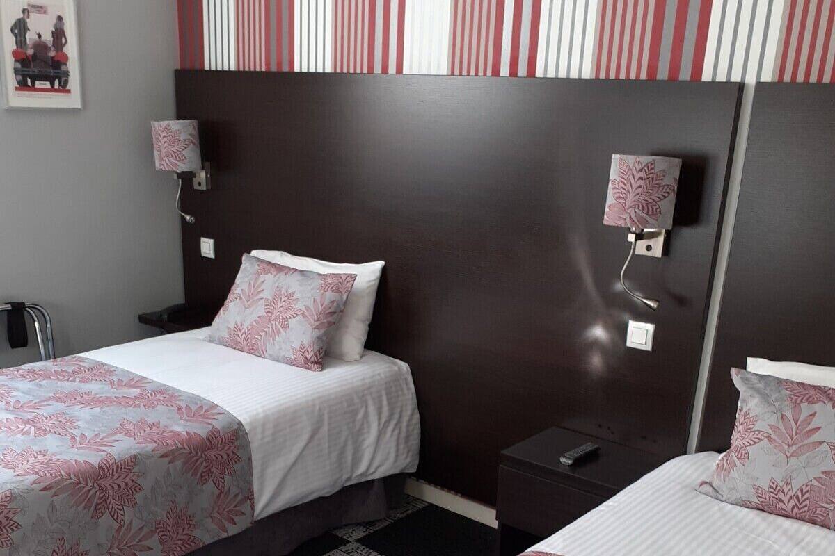 A hotel room with two beds, each with a floral-patterned bedspread. There are two bedside tables, one on each bed, with lamps on them. The room has a dark headboard and a striped wall decoration above the beds.