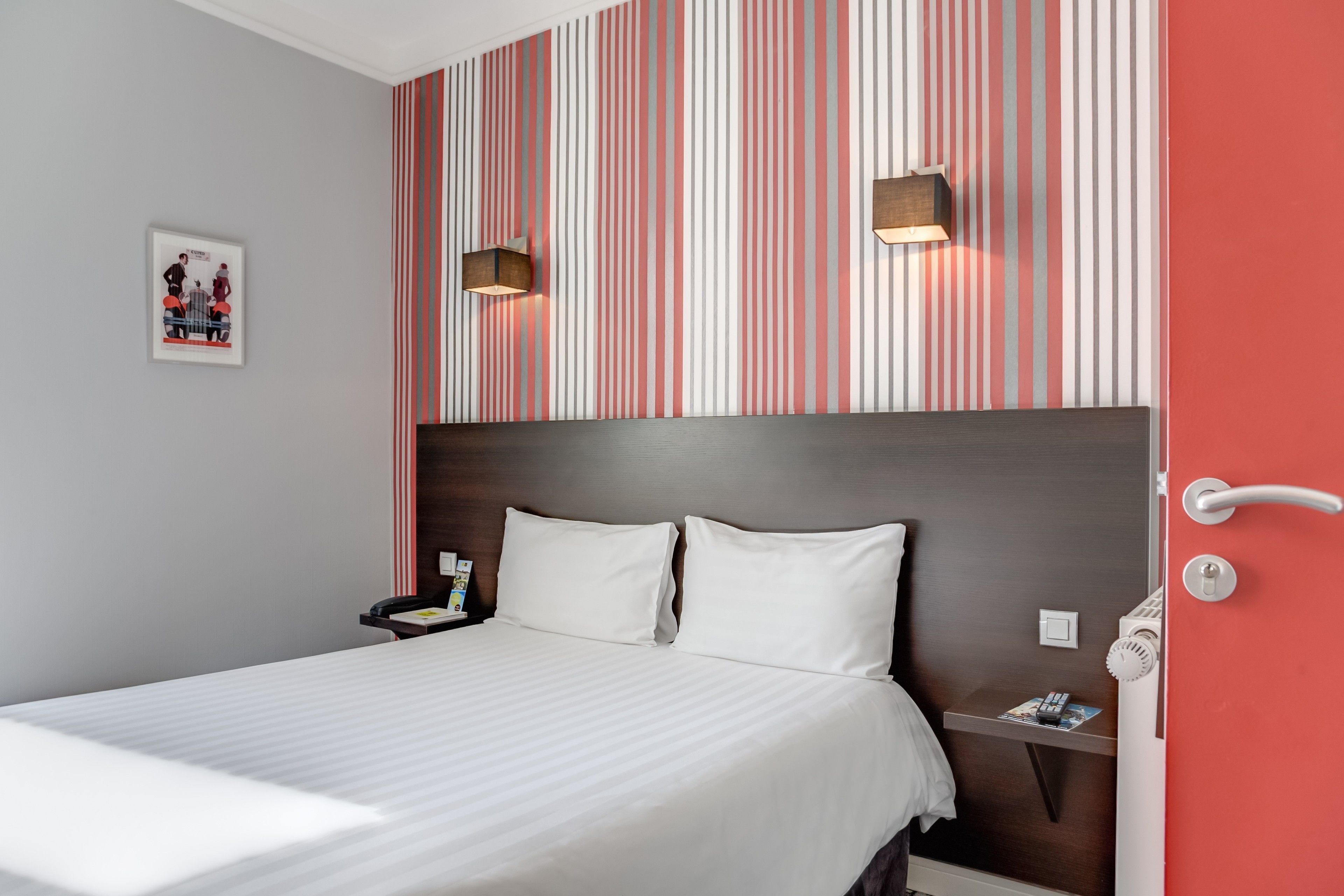 A hotel room with a striped wallpaper, a bed with white linens, a nightstand with a lamp, and a door with a handle.