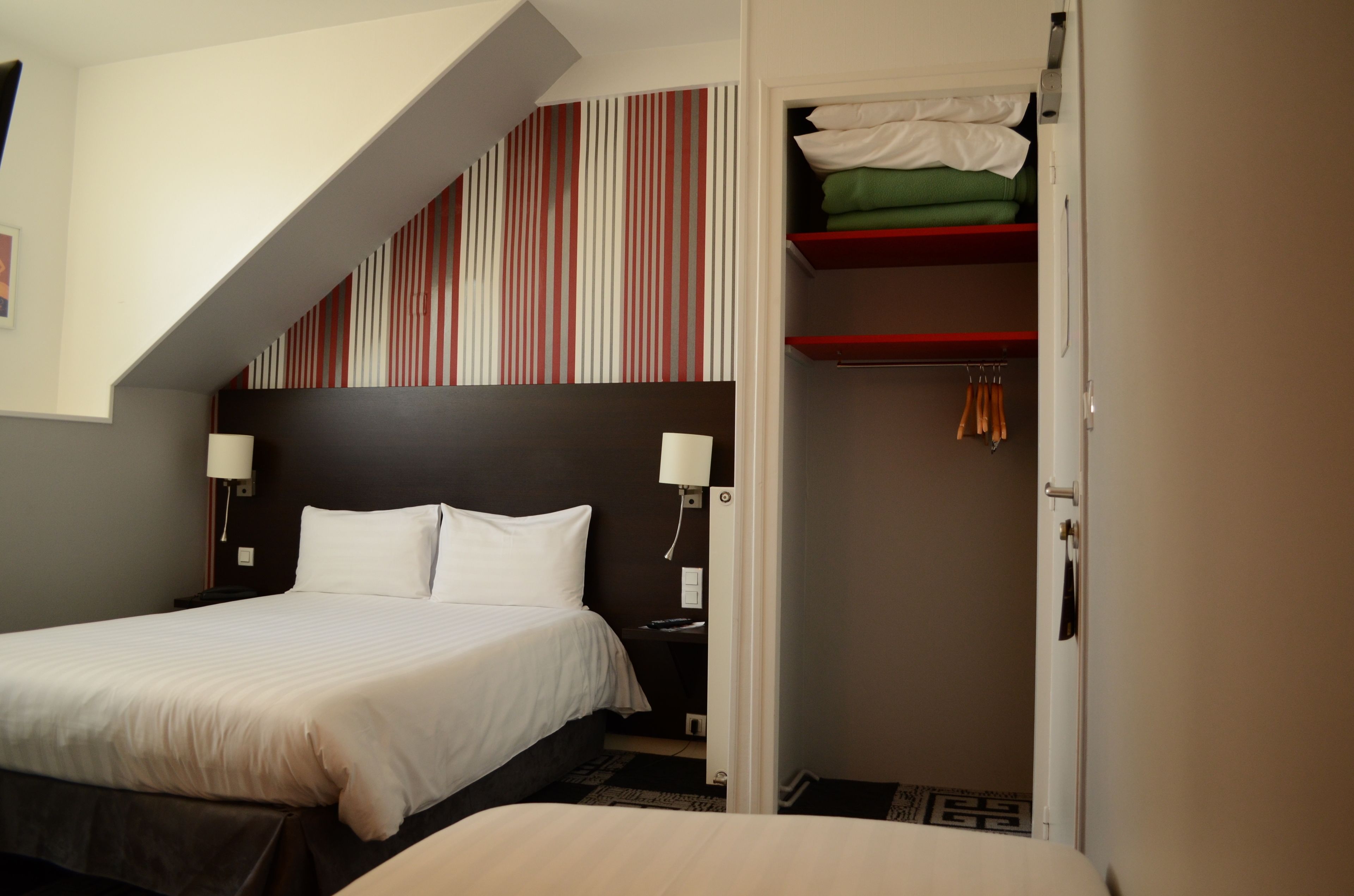 A hotel room with a bed, a nightstand, a lamp, a closet with hanging clothes, and a striped wall above the bed.
