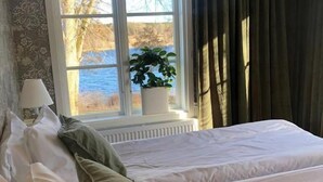 Un dormitorio con cama, almohadas, una ventana que da a vistas de árboles y un cuerpo de agua, y una planta en maceta en el alféizar.