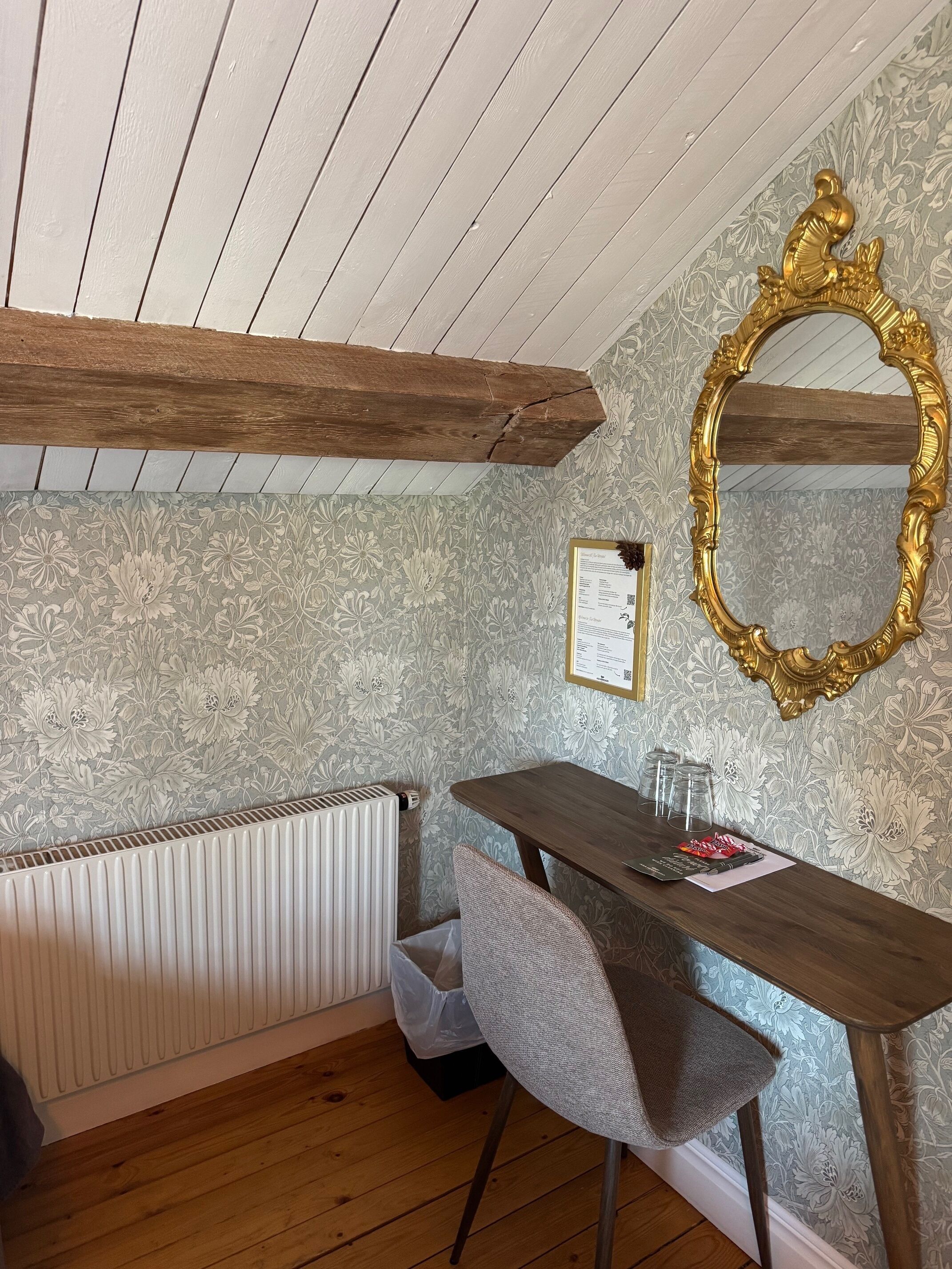 A room with a wooden ceiling, a radiator, a table with chairs, a gold-framed mirror, and patterned wallpaper.