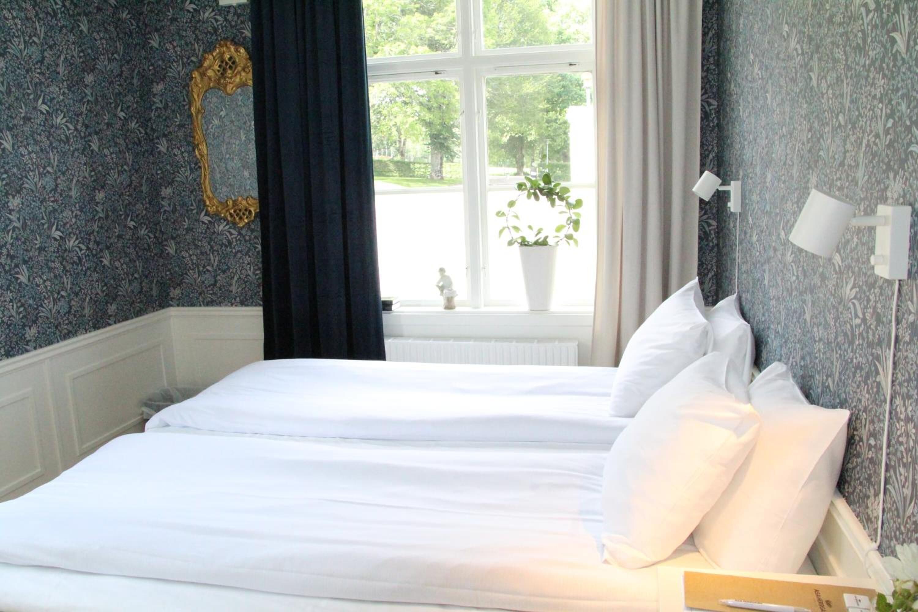A bedroom with a bed, a window with curtains, a potted plant, and a wall-mounted lamp.