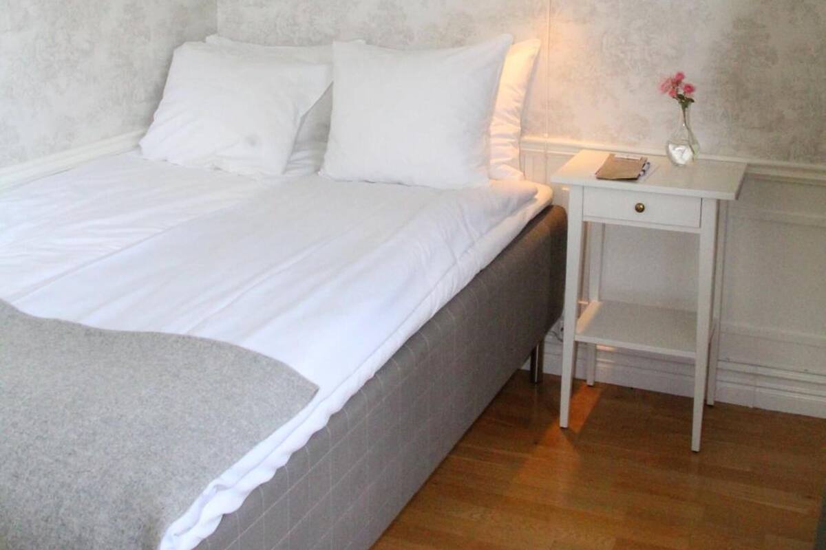 A neatly made bed with white linens, a bedside table with a vase of flowers, and a wall with a subtle floral pattern.