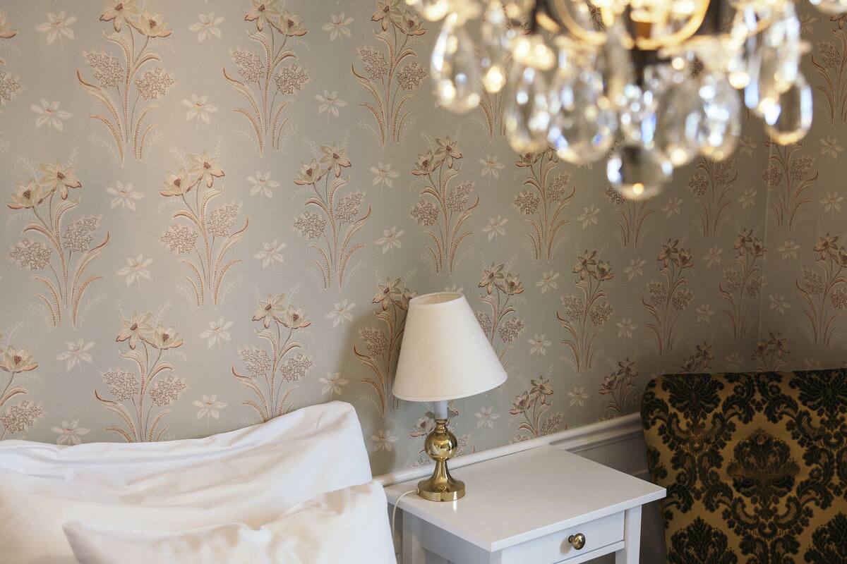 A neatly made bed with white linens and a red pillow, a white bedside table with a lamp, and a patterned armchair.