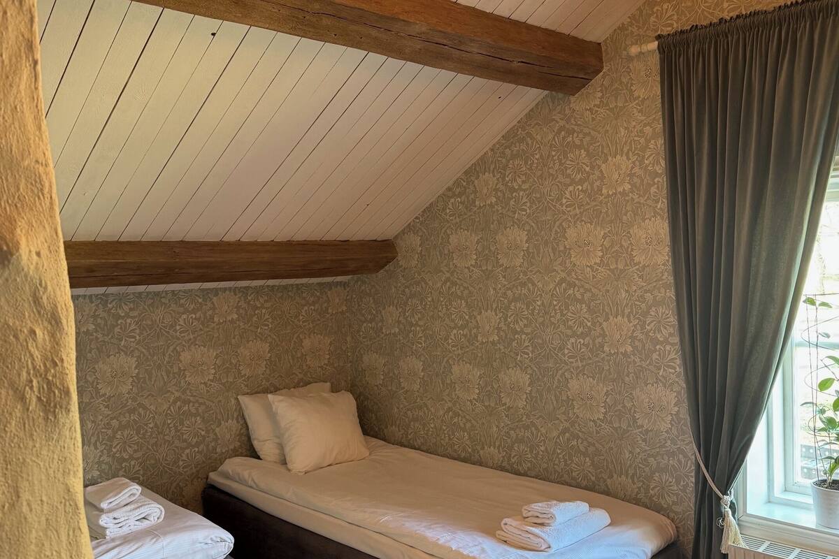 A cozy bedroom with a sloped ceiling, wooden beams, a chandelier, a bed with a grey bedspread, a bedside table, and a window with curtains.