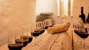 Una mesa de madera con copas de vino, una hogaza de pan, velas y una botella de vino al fondo.