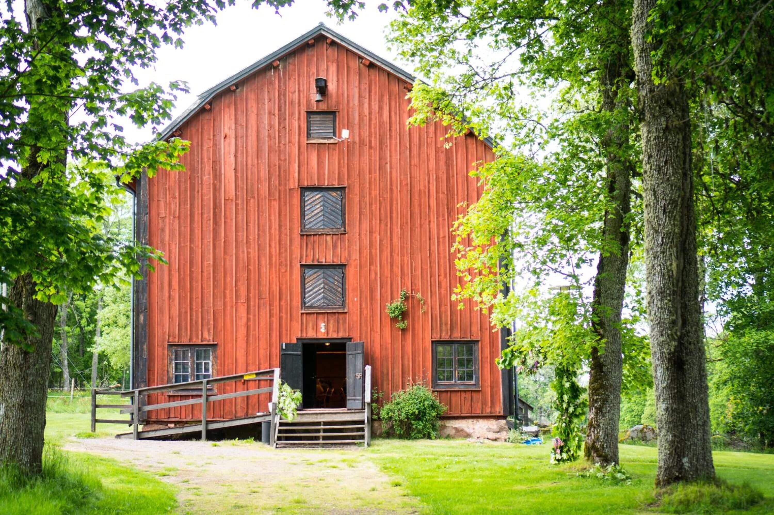 Un granero de madera roja con una puerta central y ventanas, rodeado de árboles y un área cubierta de césped.