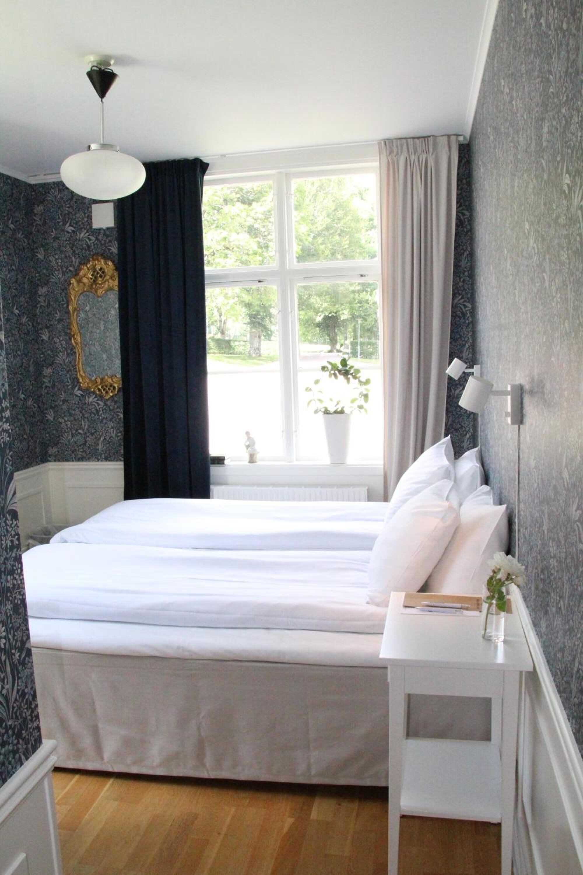 A bedroom with a large bed, a bedside table, a window with curtains, and a wall with a decorative mirror.