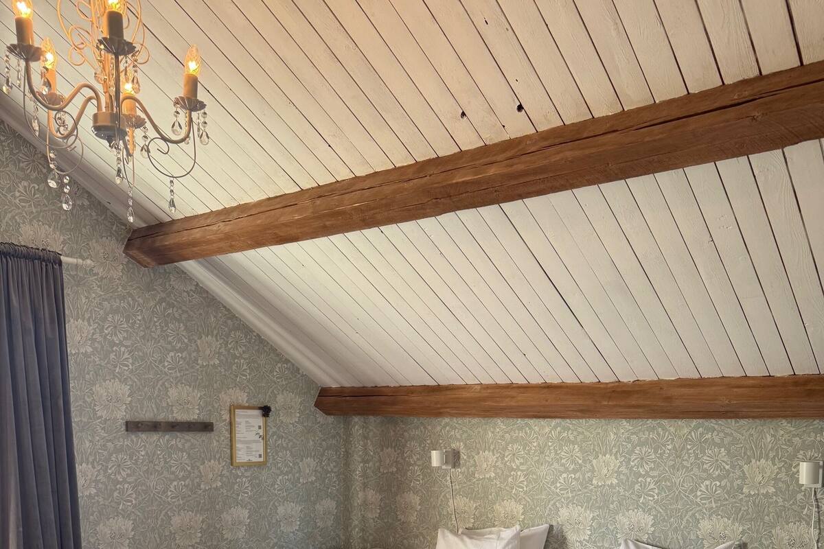 A bedroom with two beds, wooden beams, a chandelier, and a floral wallpaper pattern.
