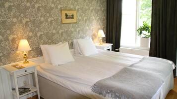 Una cama bien hecha con sábanas blancas, dos mesitas de noche con lámparas, un cuadro enmarcado en la pared y una ventana con cortinas.