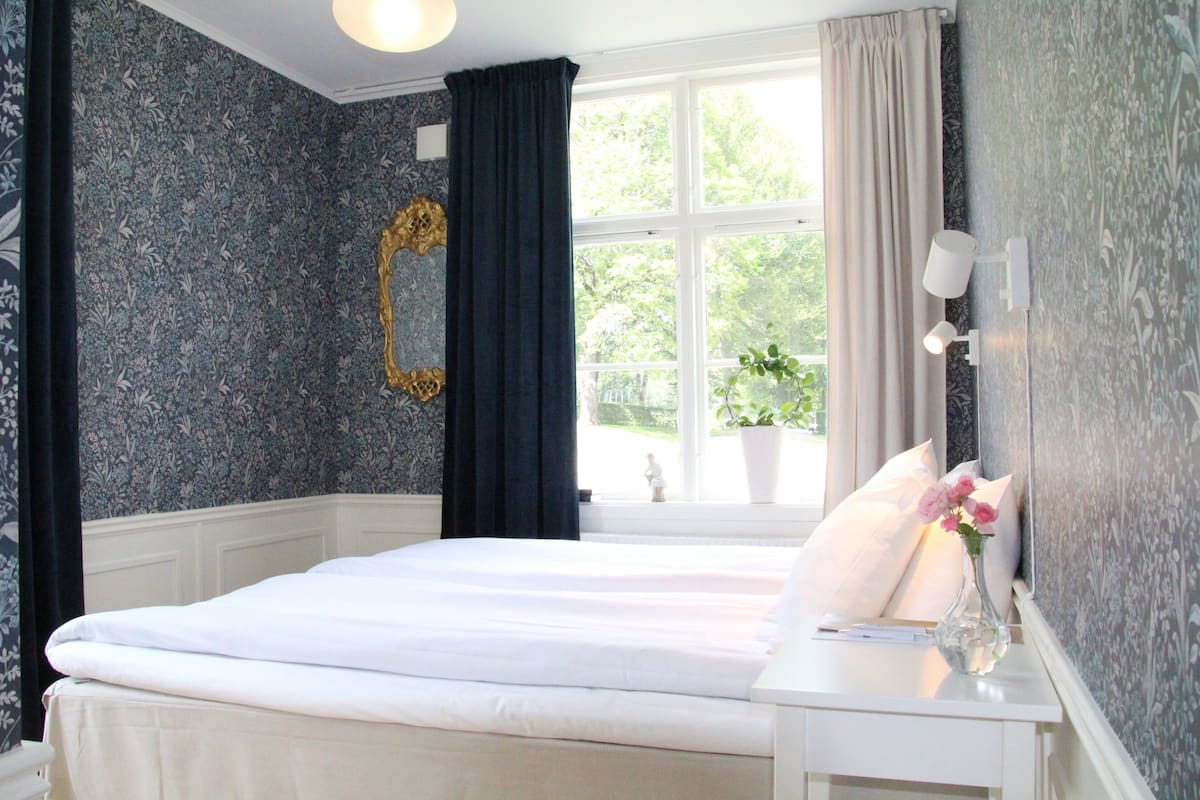 A bedroom with a large bed, a bedside table with a vase of flowers, a window with curtains, and a wall with a decorative mirror.