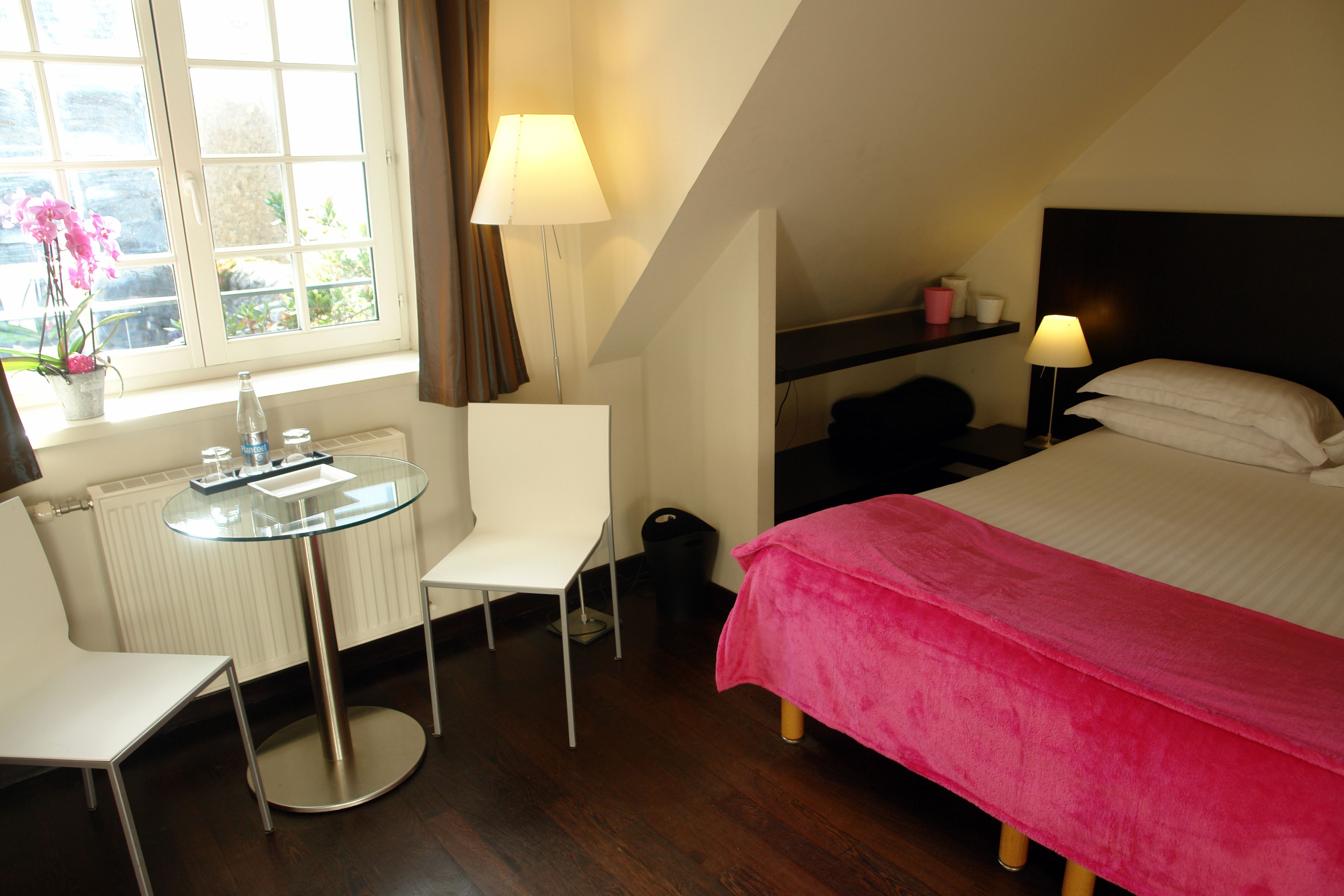Ein Hotelzimmer mit Bett, Sessel, kleinem Tisch, Lampe, Fenster mit Vorhängen und Heizkörper.