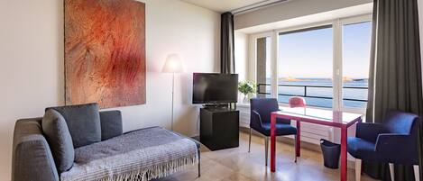 A modern living room with a sofa, TV, and dining area with a view of the sea.