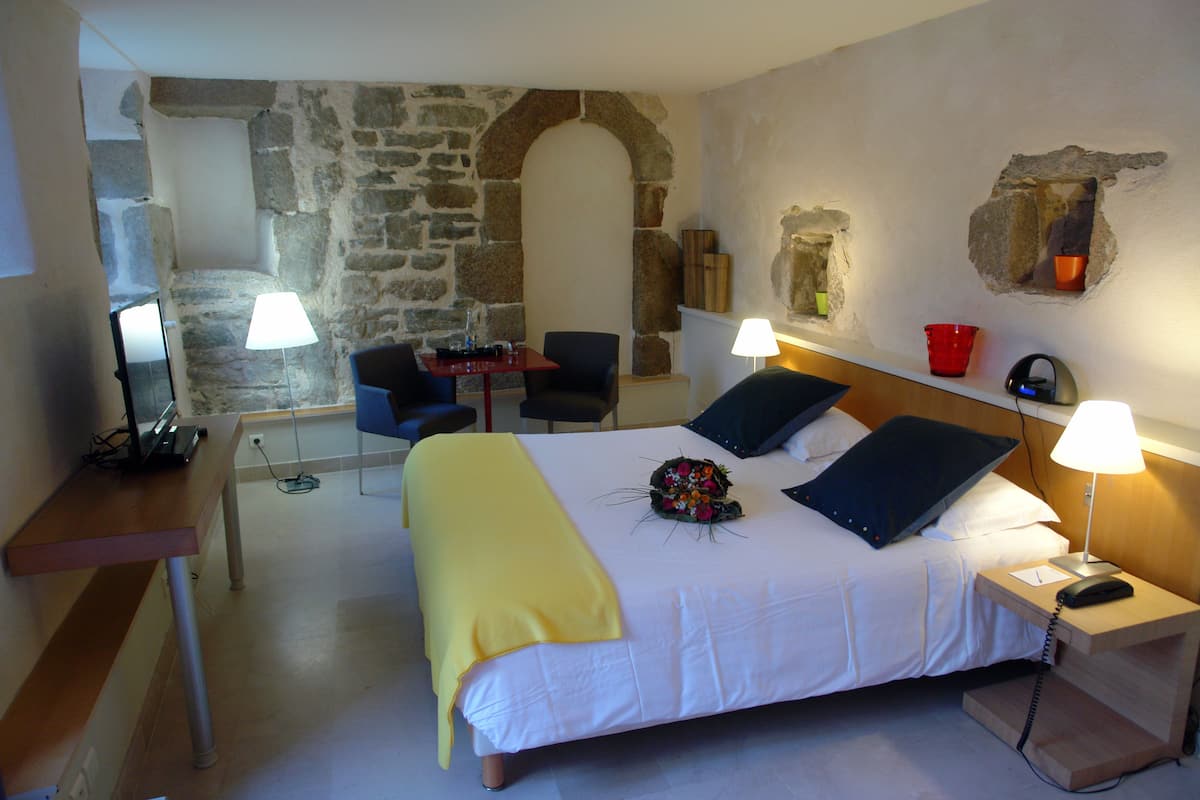 A hotel room with a stone wall, a bed with white bedding, a desk with a lamp, a chair, and a nightstand with a phone.