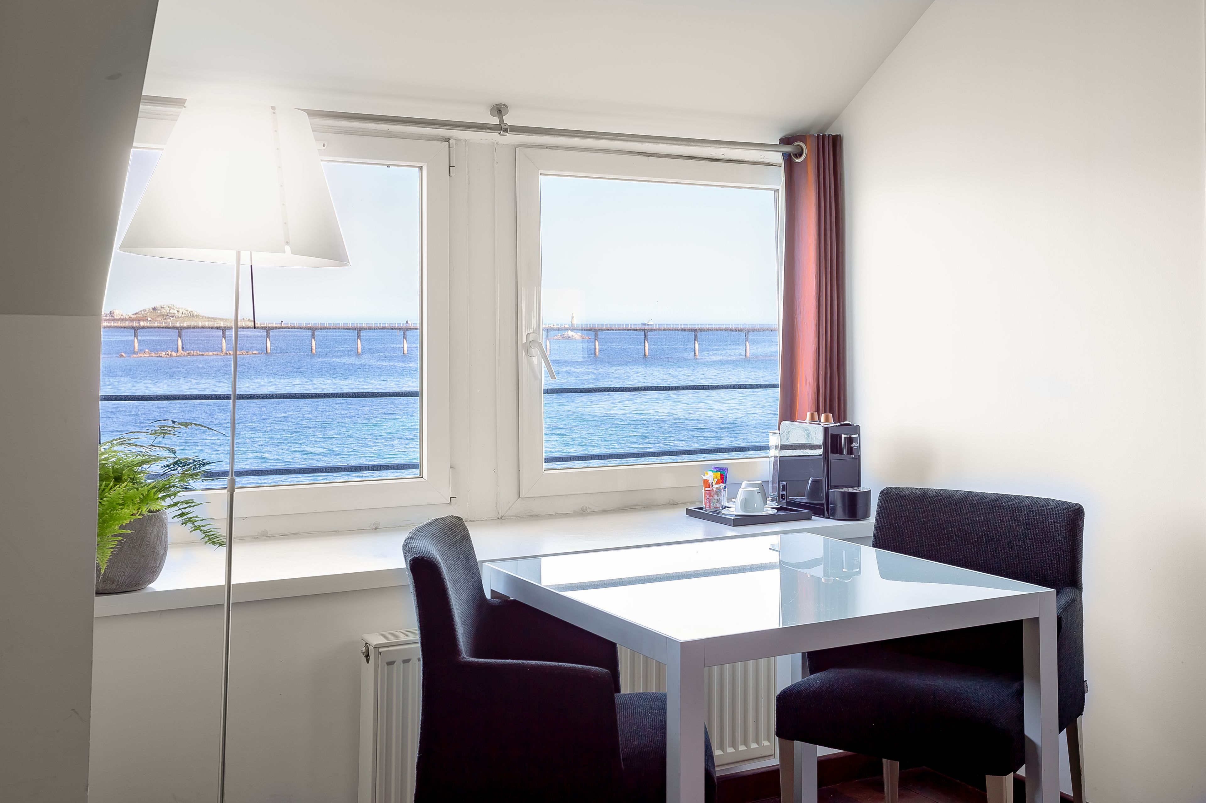 Une pièce avec une table blanche, deux chaises noires, une plante en pot, une lampe, et une vue sur la mer et un pont.