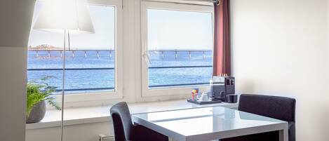 Ein Zimmer mit einem weißen Tisch, zwei schwarzen Stühlen, einer Topfpflanze, einer Lampe und Blick auf das Meer und eine Brücke.