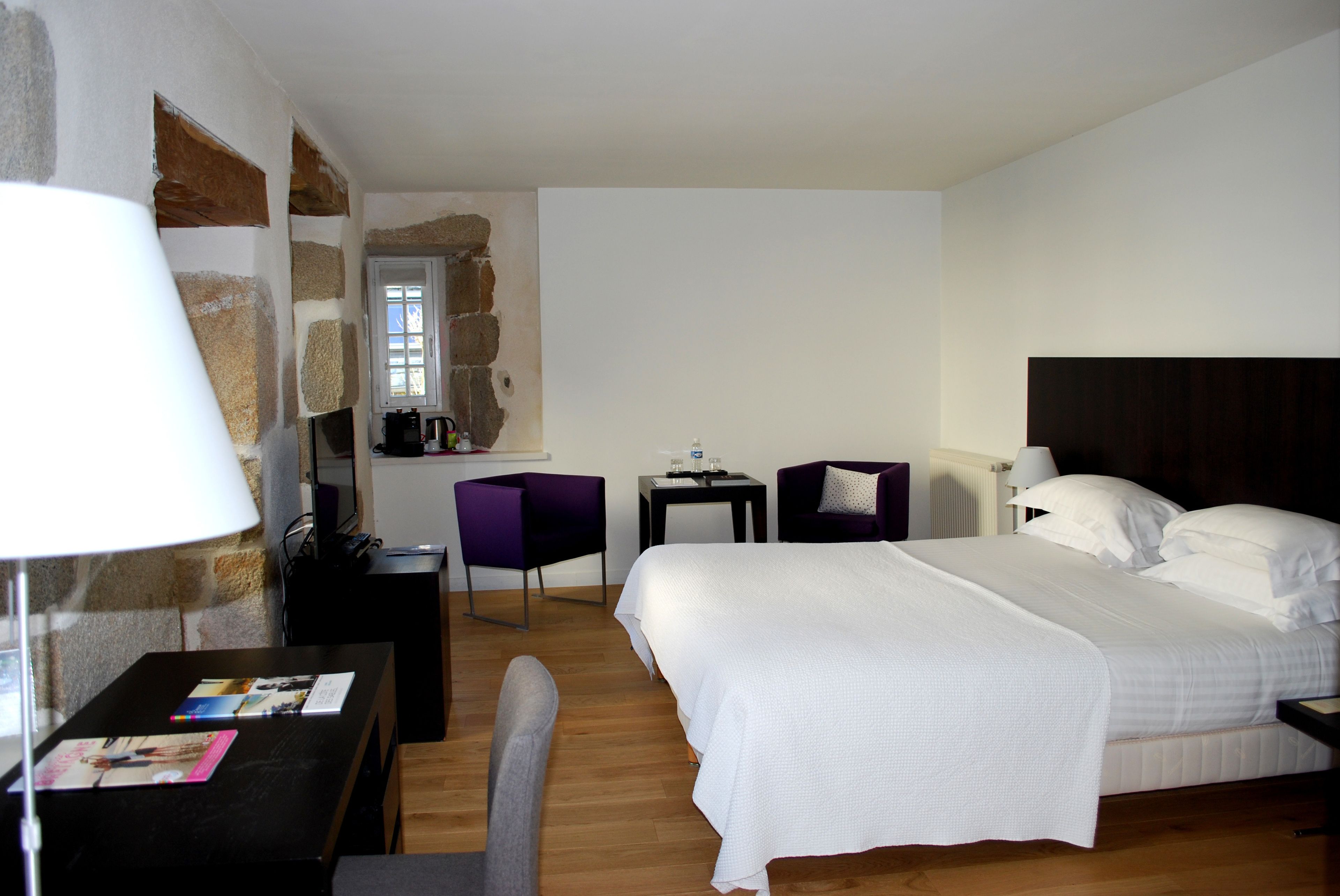 A hotel room with a bed, a desk with a lamp, a chair, a small table, and a window.