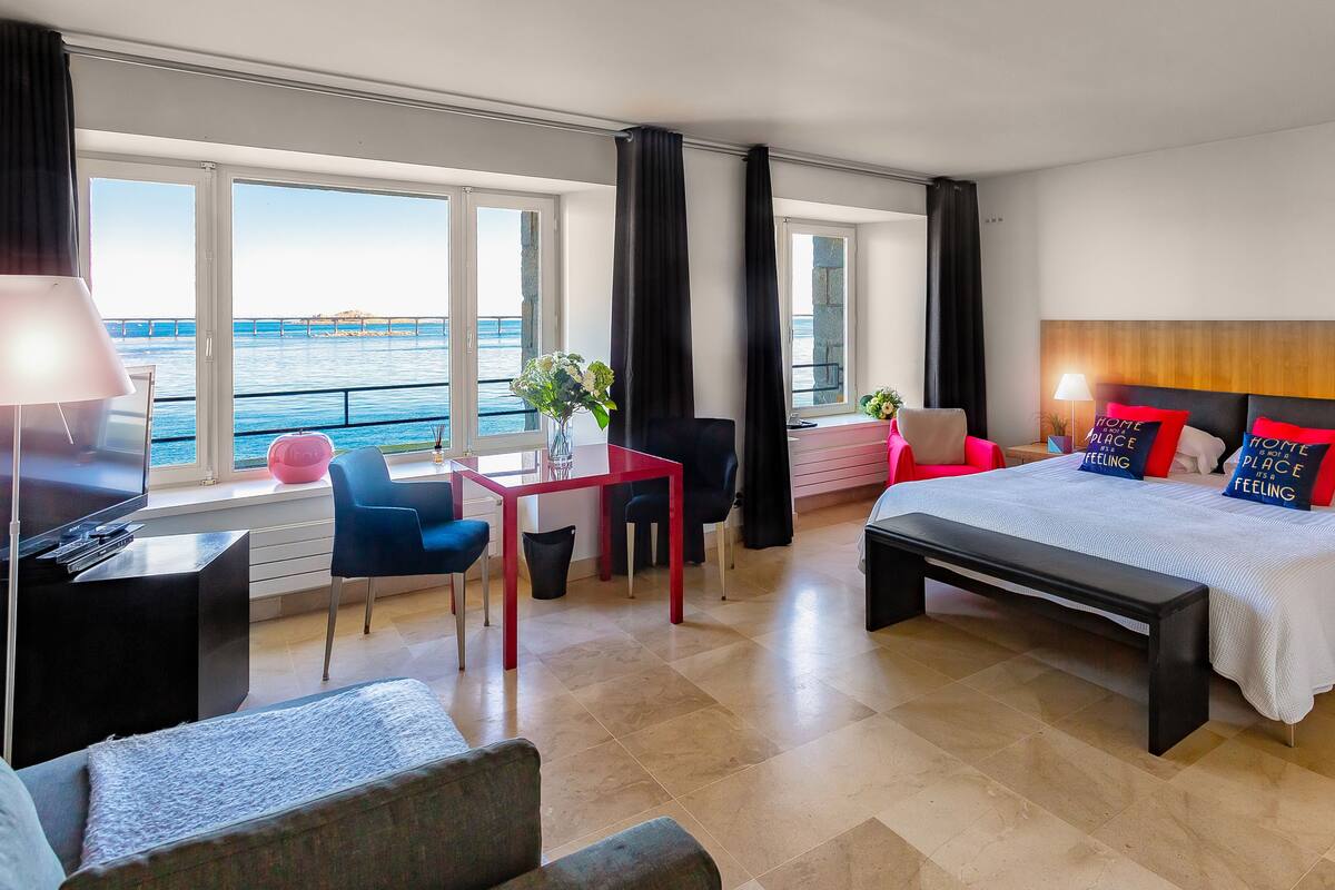 A hotel room with a bed, bedside tables, a desk, and a chair. There are large windows with curtains, a view of the sea, and a potted plant.