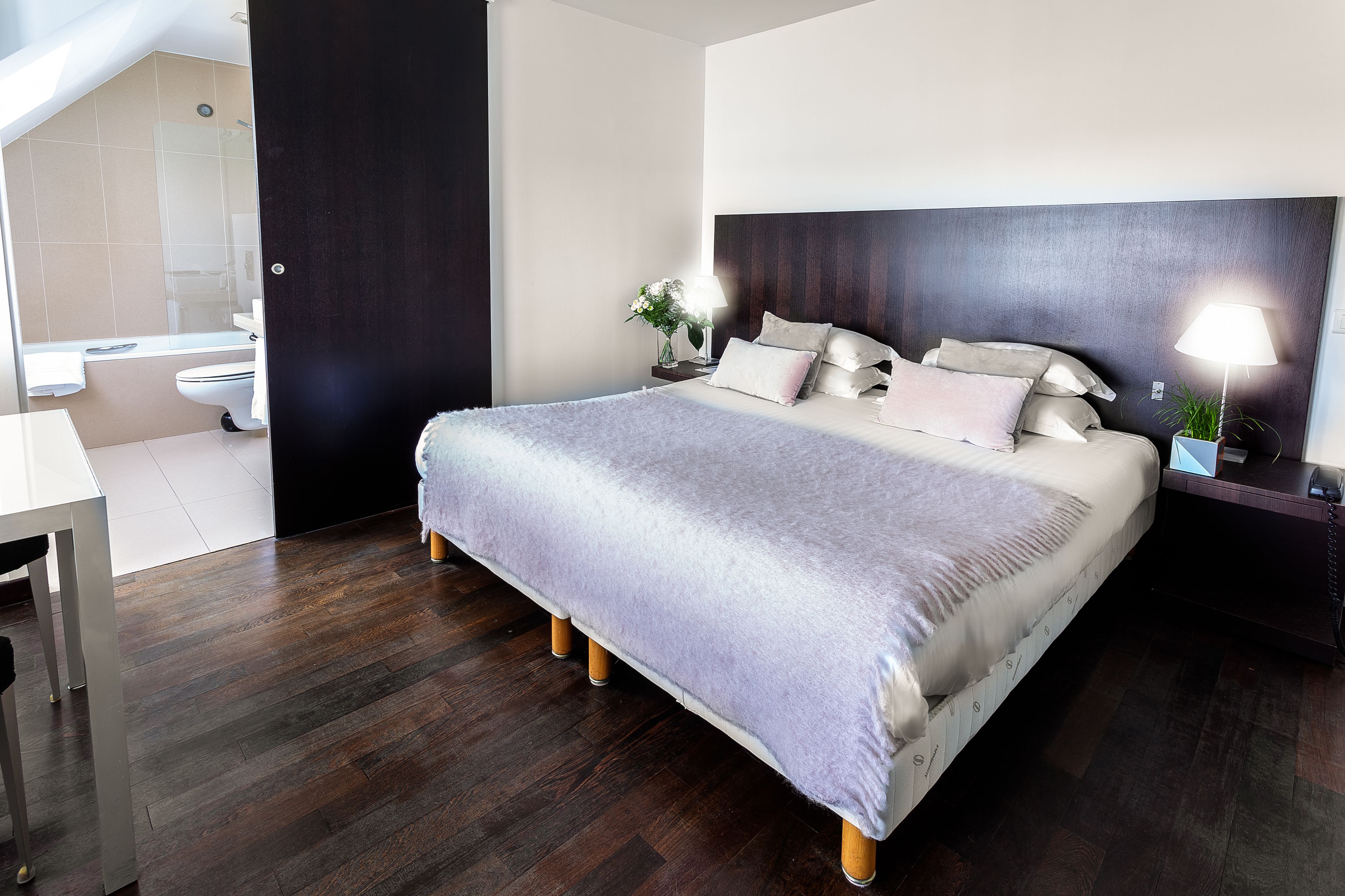 Een moderne slaapkamer met een groot bed, nachtkastjes met lampen, een donker hoofdeinde en uitzicht op de badkamer.