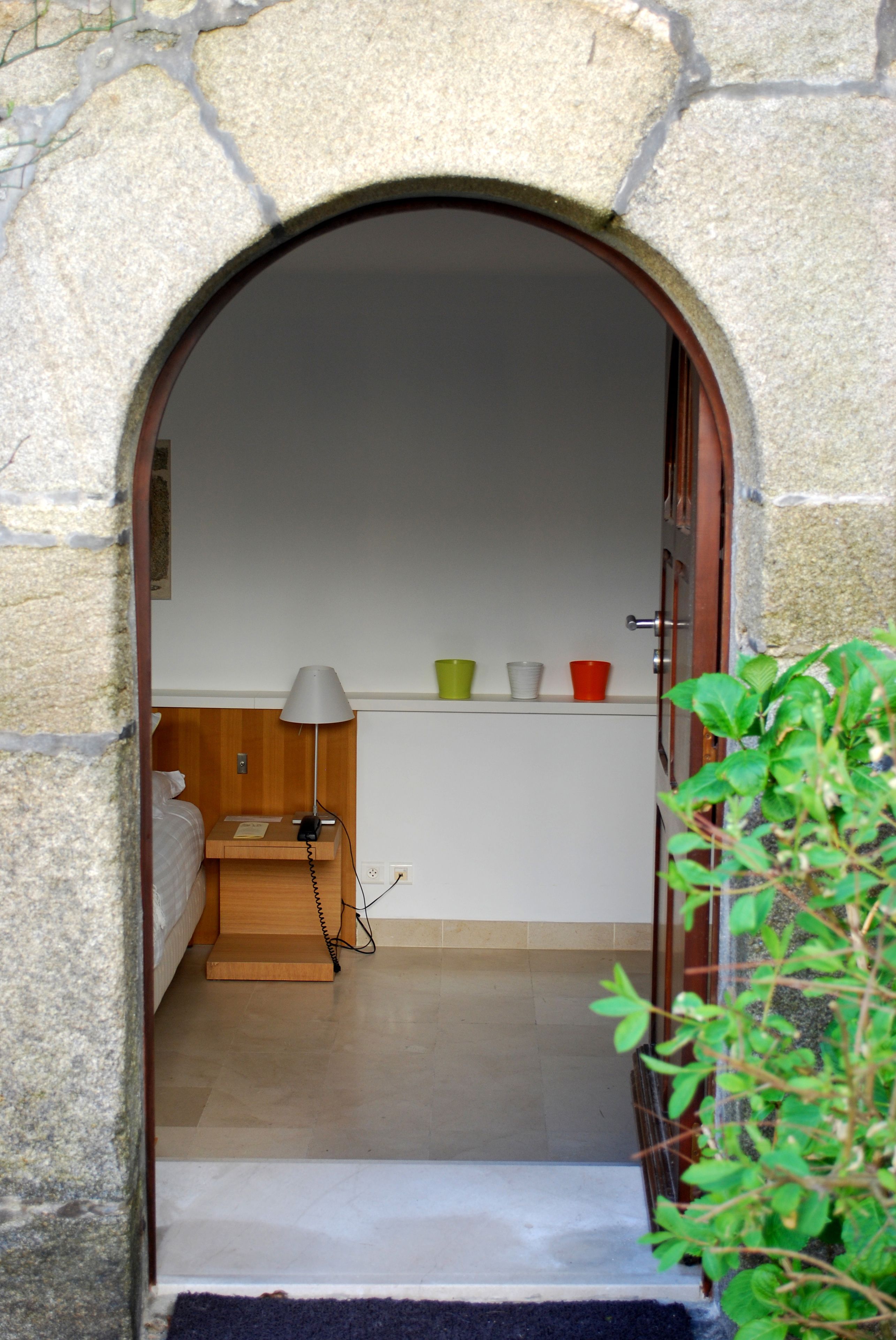 Un arc en pierre mène à une petite pièce où se trouvent un lit, un bureau, une lampe et une étagère avec des tasses.