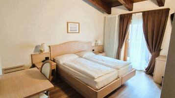 A double bed with a wooden headboard, a bedside table with a lamp, a desk with a chair, and a window with curtains.