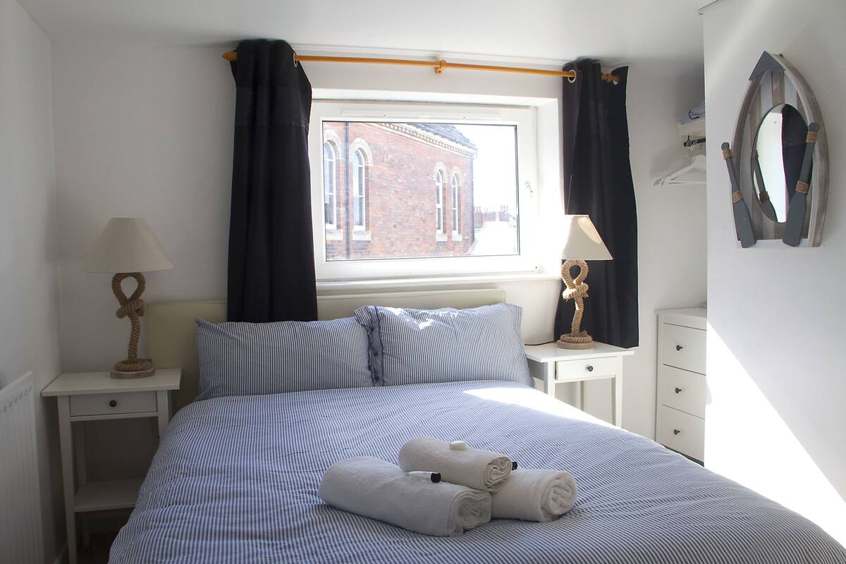 A neatly made bed with striped bedding, two bedside tables with lamps, a window with curtains, and a mirror on the wall.