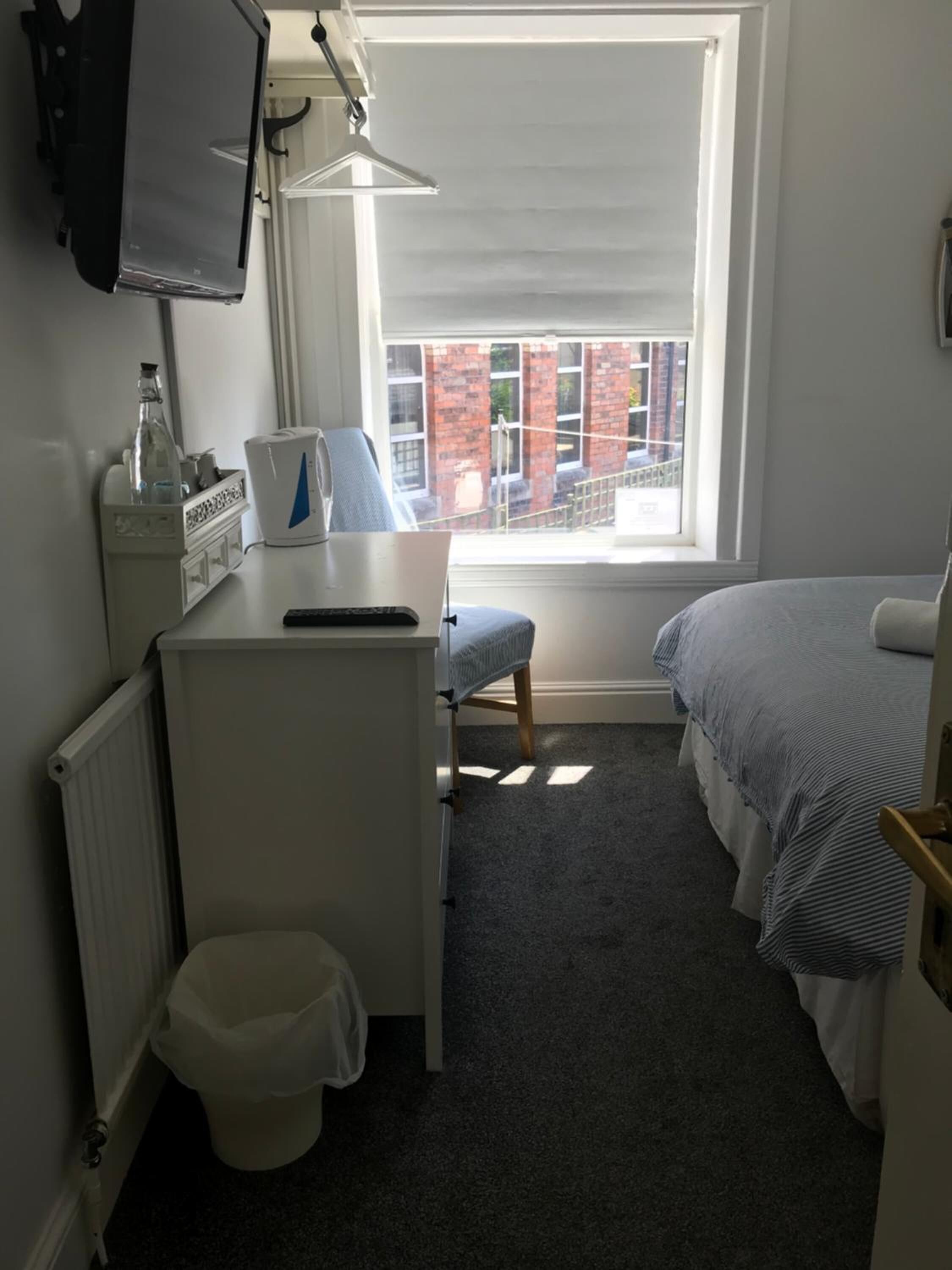 Et lille, godt oplyst værelse med en enkelt seng, et skrivebord med stol, en radiator, et vindue med udsigt til bygninger og et fjernsyn ophængt på væggen.