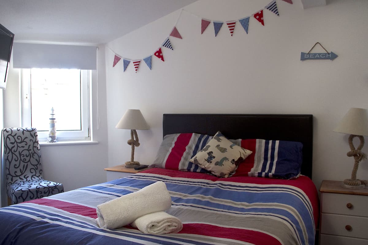 A neatly made bed with striped bedding, a decorative pillow, and a rolled towel. There are two bedside lamps, a chair with a patterned cushion, and a wall-mounted sign indicating the direction to the beach.