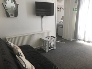 A living room with a black sofa, a small white side table with a book, a flat-screen TV mounted on the wall, and a white radiator. - Timrick Lodge Hotel (Weymouth)