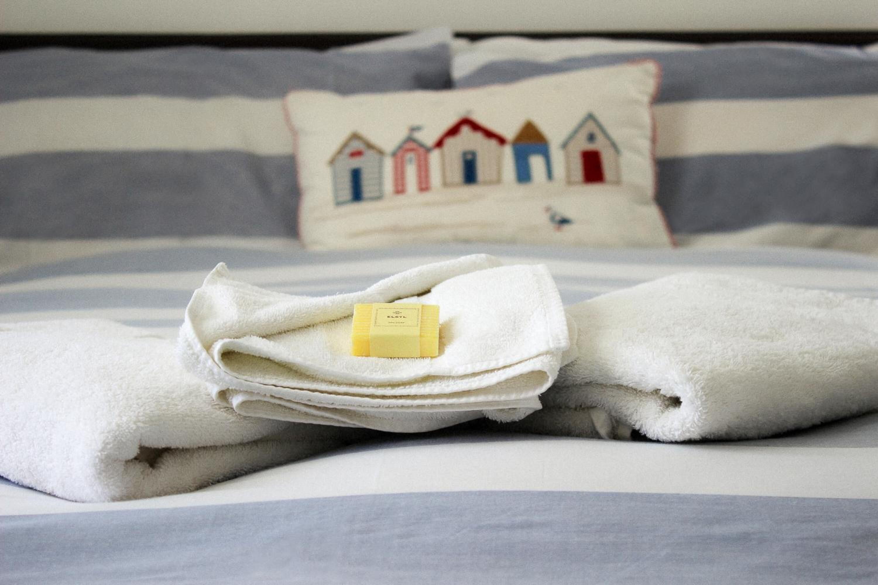 Una toalla blanca doblada con cuidado, una barra de jabón sobre una cama con ropa de cama a rayas y un almohadón con diseño de casa.