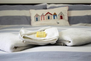 Una toalla blanca doblada con cuidado, una barra de jabón sobre una cama con ropa de cama a rayas y un almohadón con diseño de casa.
