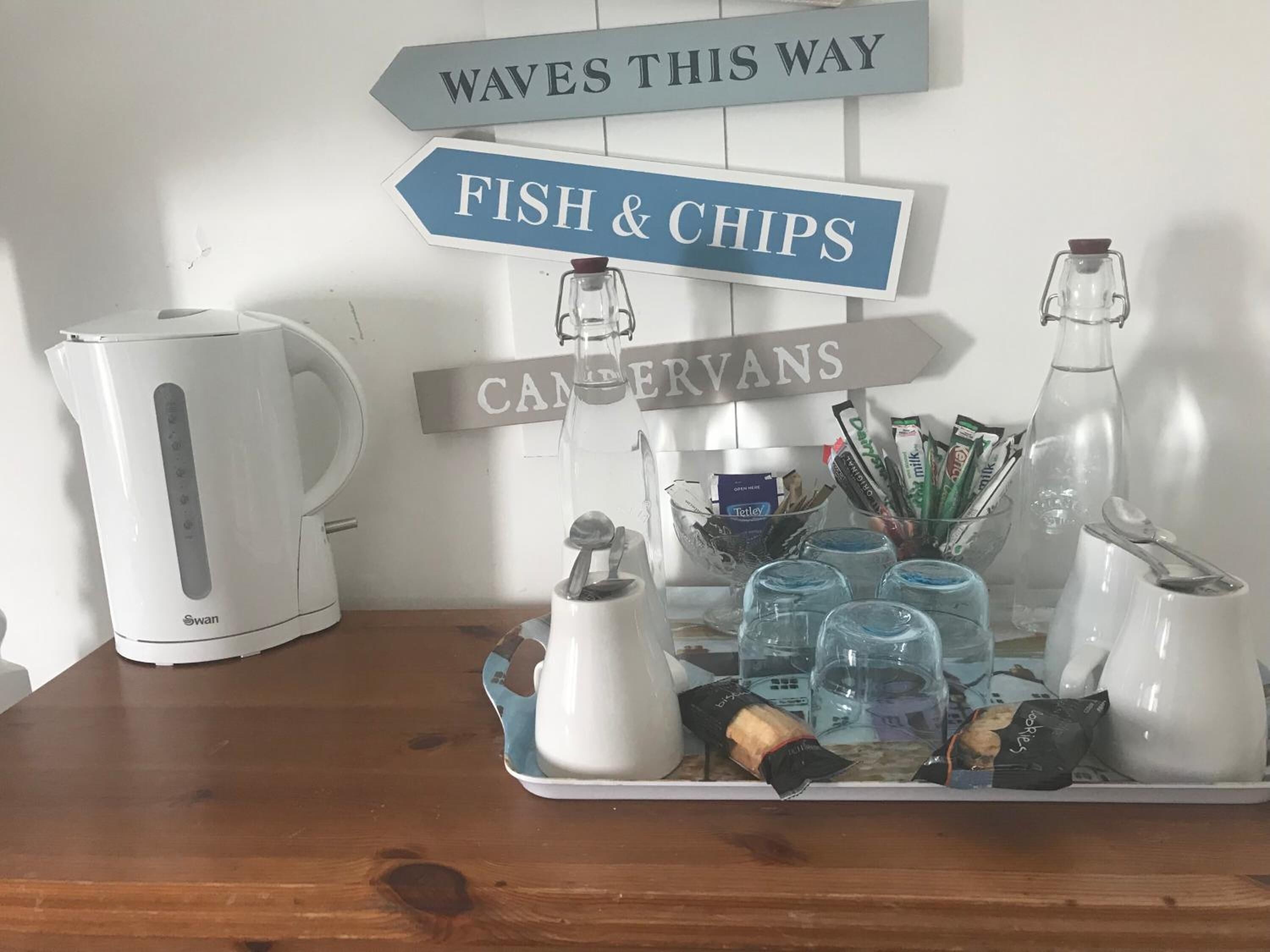 Una encimera de cocina con una tetera, una bandeja de condimentos y letreros que indican 'ESTE ES EL CAMINO A WAVES', 'FISH & CHIPS' y 'CANCERVANS'.