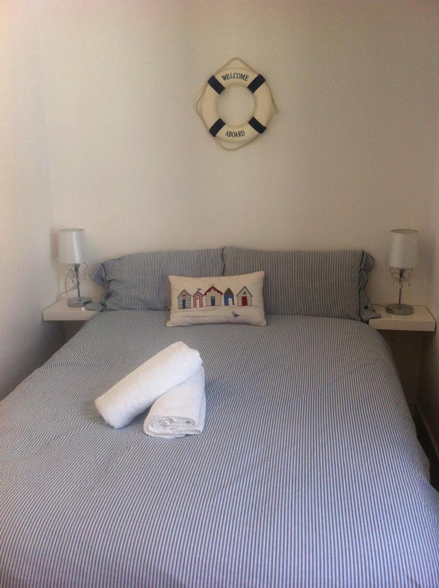 A neatly made bed with striped bedding, a folded towel, a pillow with a house design, and a welcome aboard life preserver on the wall.