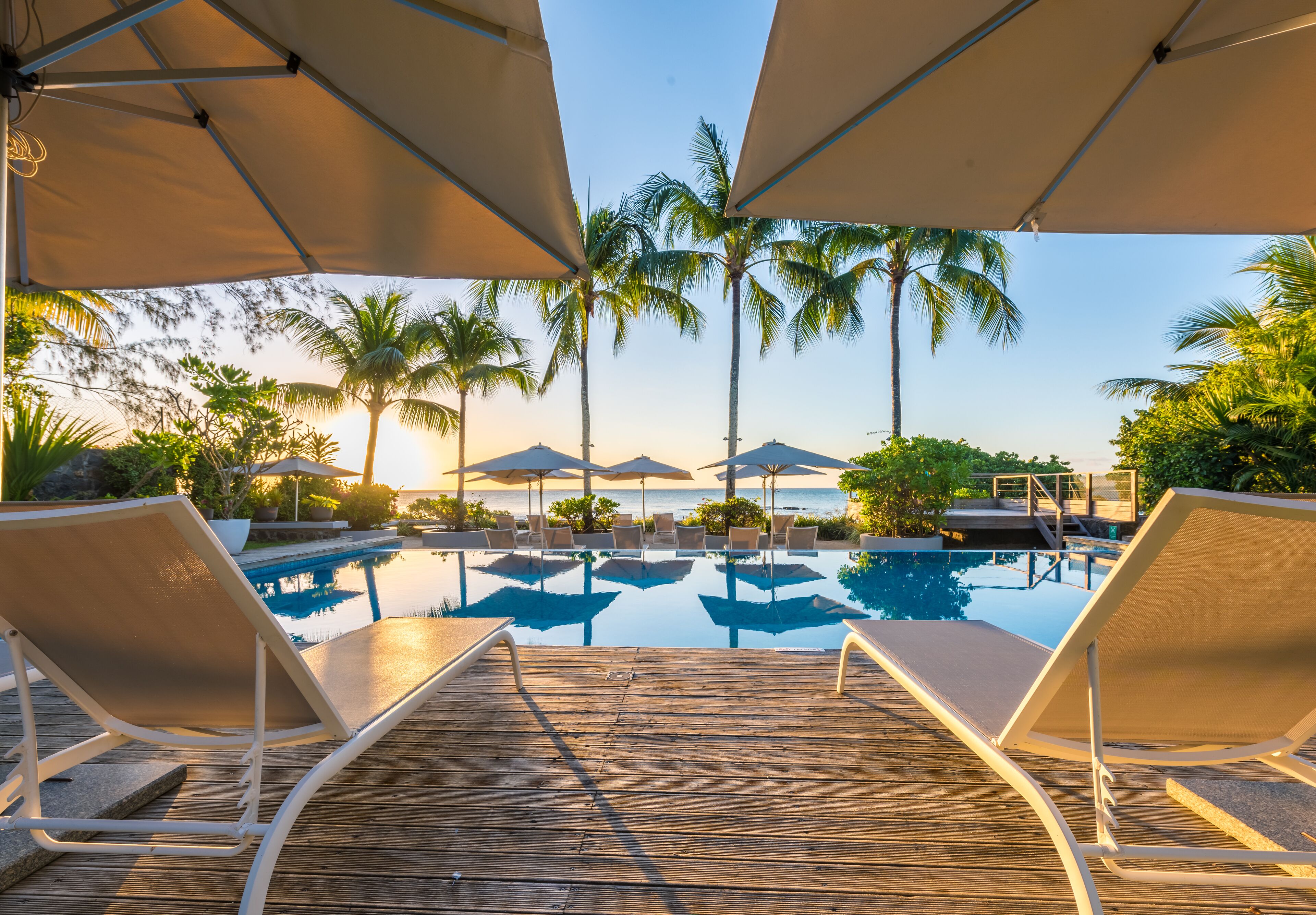 A wooden deck with lounge chairs and umbrellas overlooking a pool with palm trees and a view of the ocean.