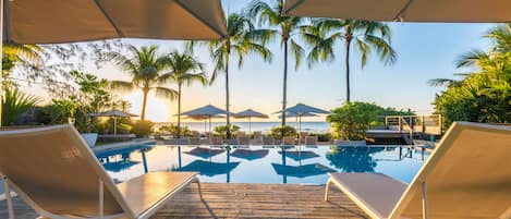 A wooden deck with lounge chairs and umbrellas overlooking a pool with palm trees and a view of the ocean.