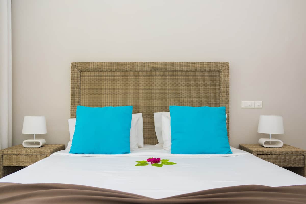 Una cama bien hecha con dos almohadas azul brillante, una colcha blanca y una sola flor morada. Hay dos mesitas de noche con lámparas blancas.